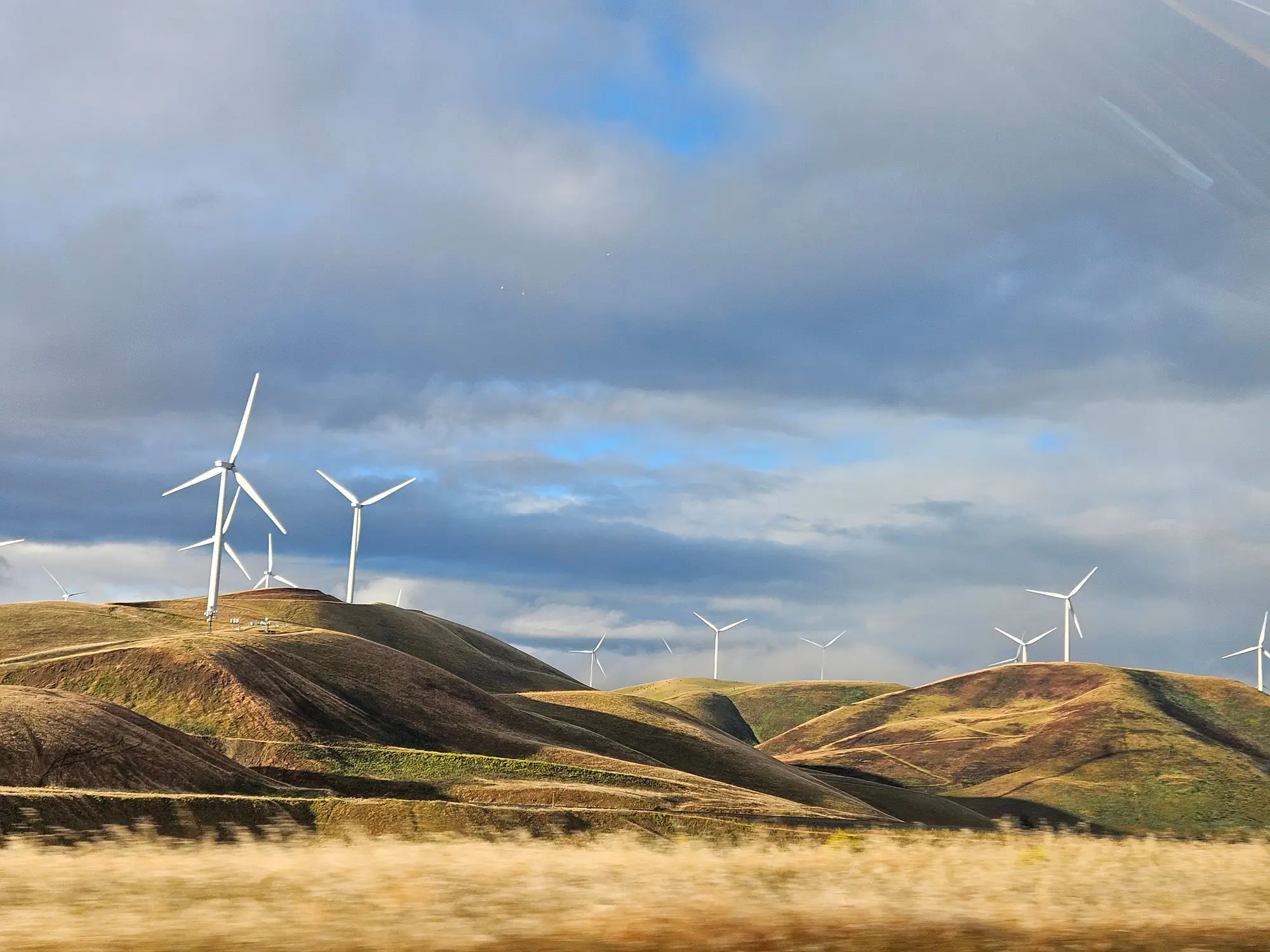 Wind turbines spaced along a ridge of dry, sunlit grassland under broken cloud